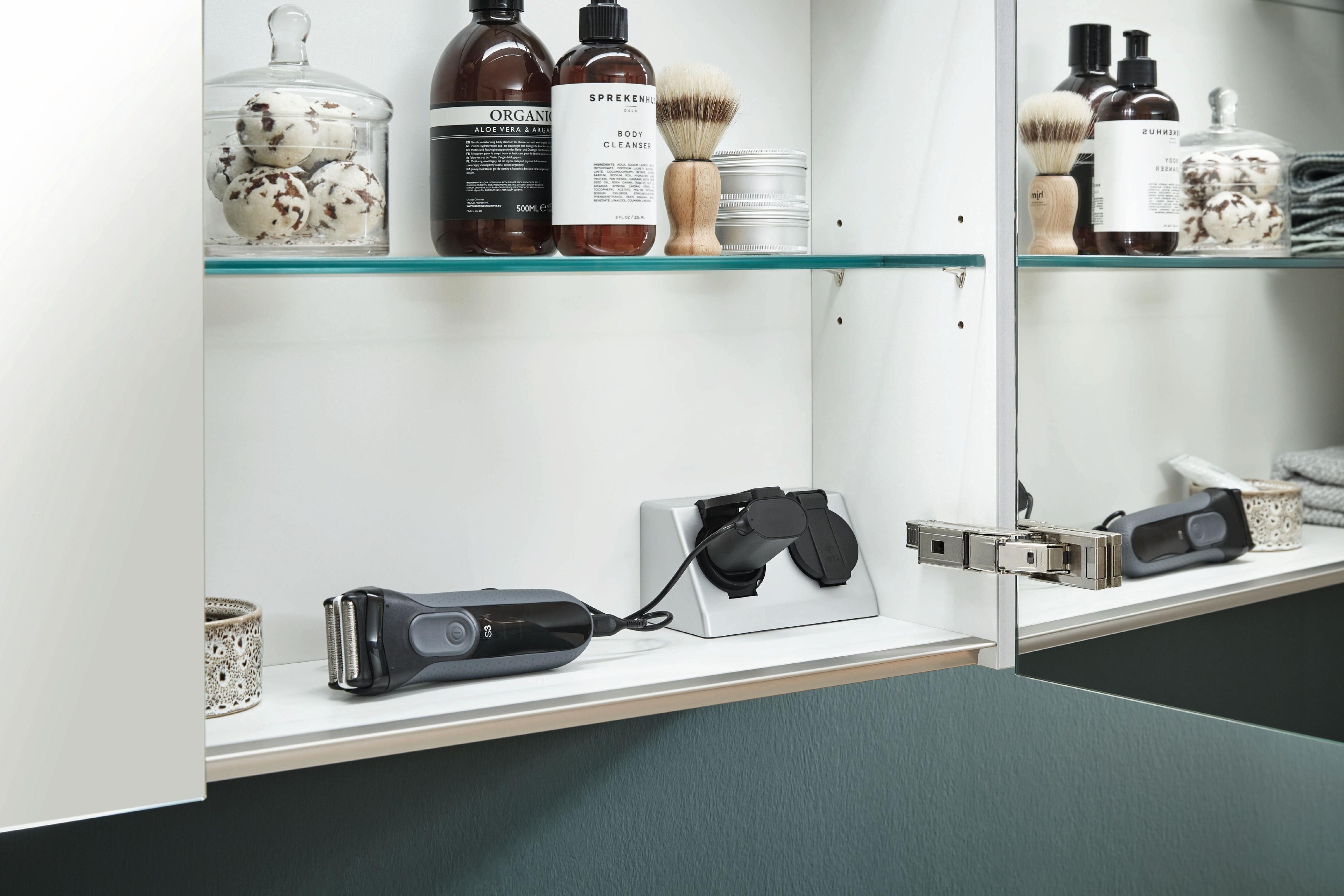 Offener Spiegelschrank mit integrierten Steckdosen und Pflegeprodukten wie Rasierer und Lotionen.
