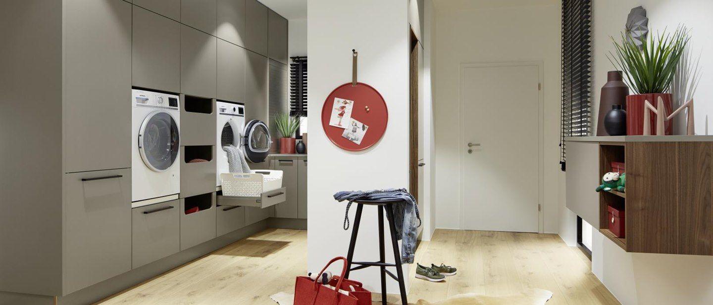 Moderner Hauswirtschaftsraum mit Einbauschränken in Taupe, integrierter Waschmaschine und Trockner, ausziehbarem Wäschekorb und dekorativen Akzenten in Rot und Holzoptik.