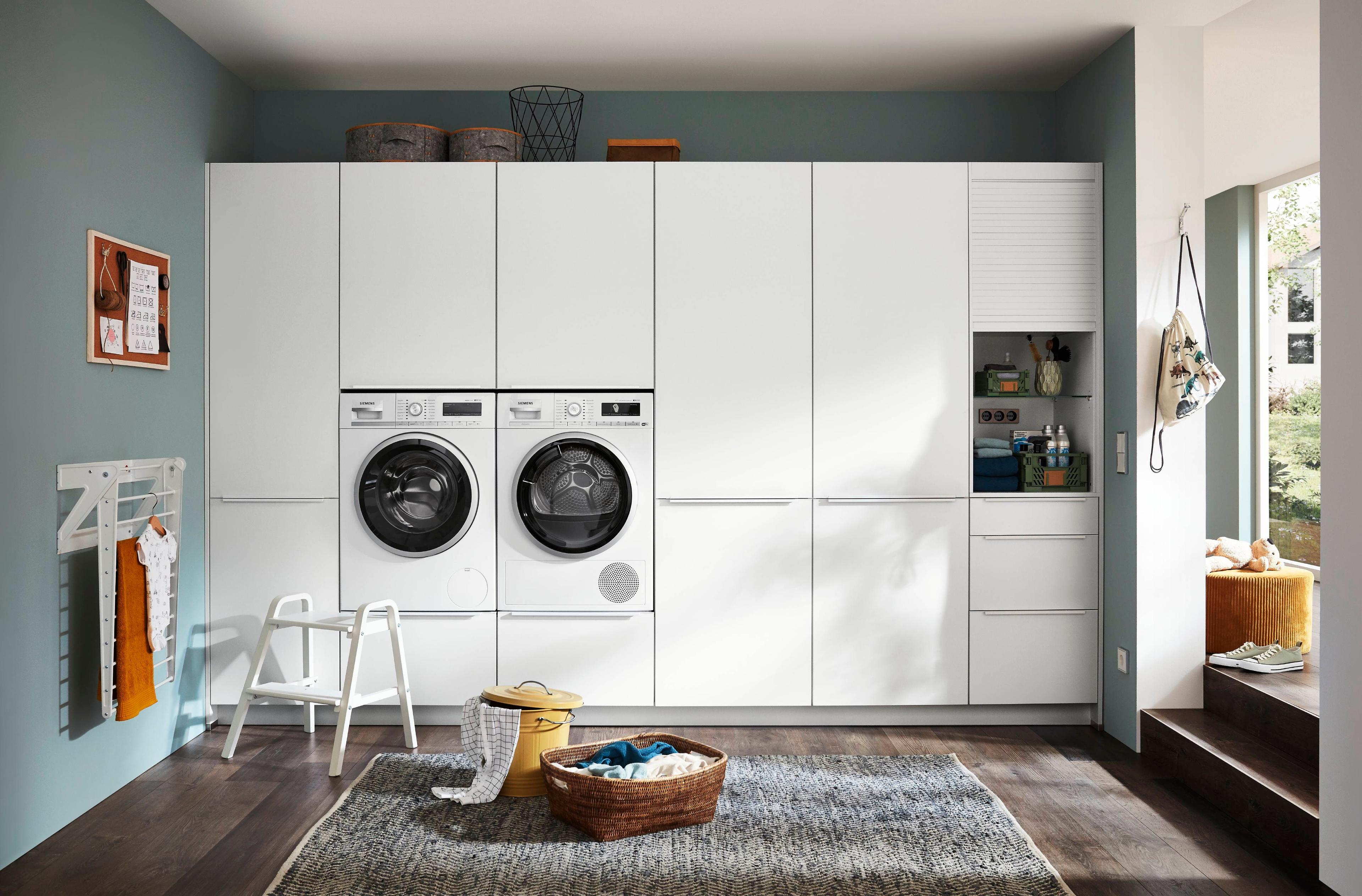 Waschküche mit weißer Schrankwand, eingebauter Waschmaschine und Trockner, hellblauen Wänden und Weidekörben für Wäsche auf dunklem Holzboden.
