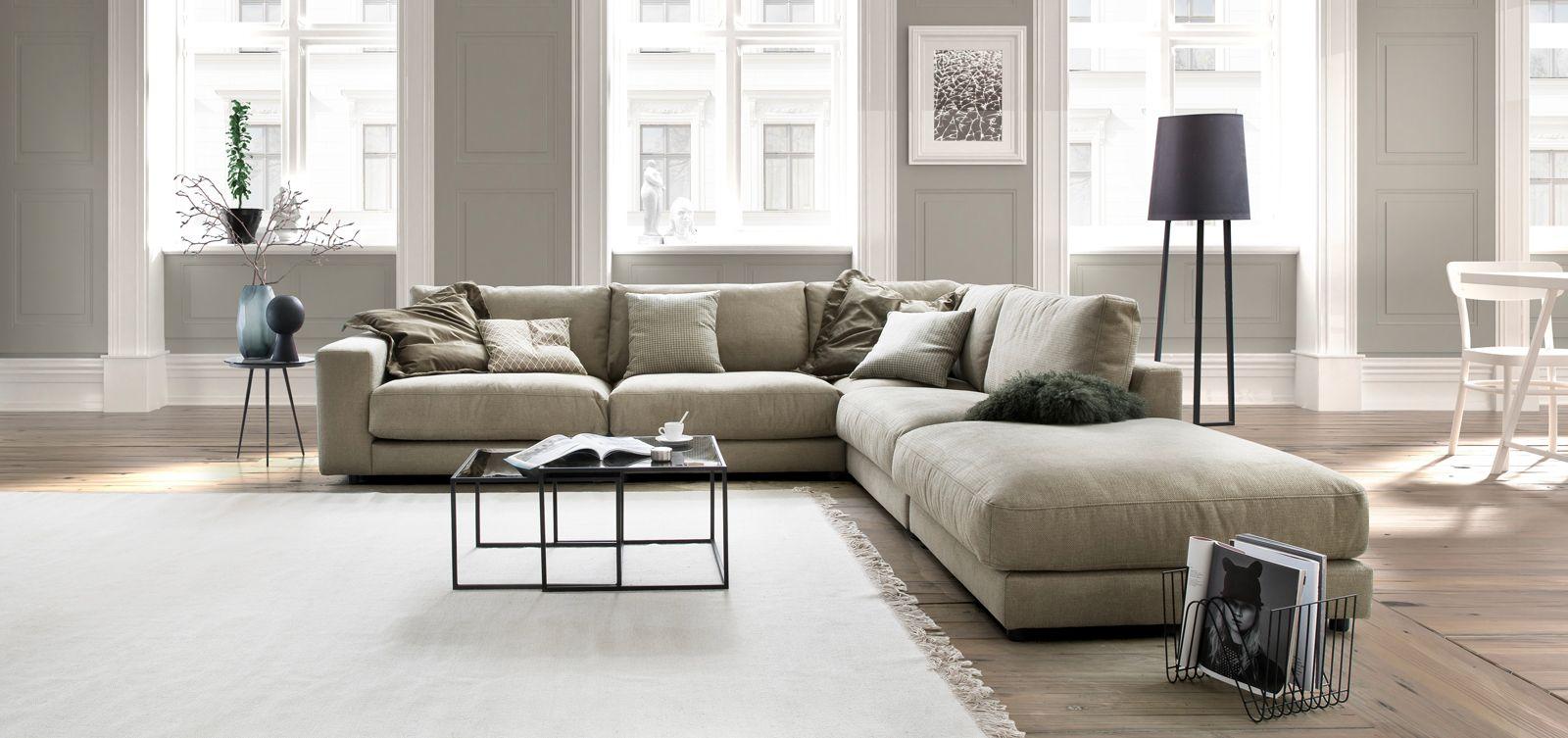 Großzügiges Ecksofa in Beige mit dekorativen Kissen in einem stilvollen, hellen Wohnraum mit Holzfußboden, hohen Fenstern und modernen Accessoires.