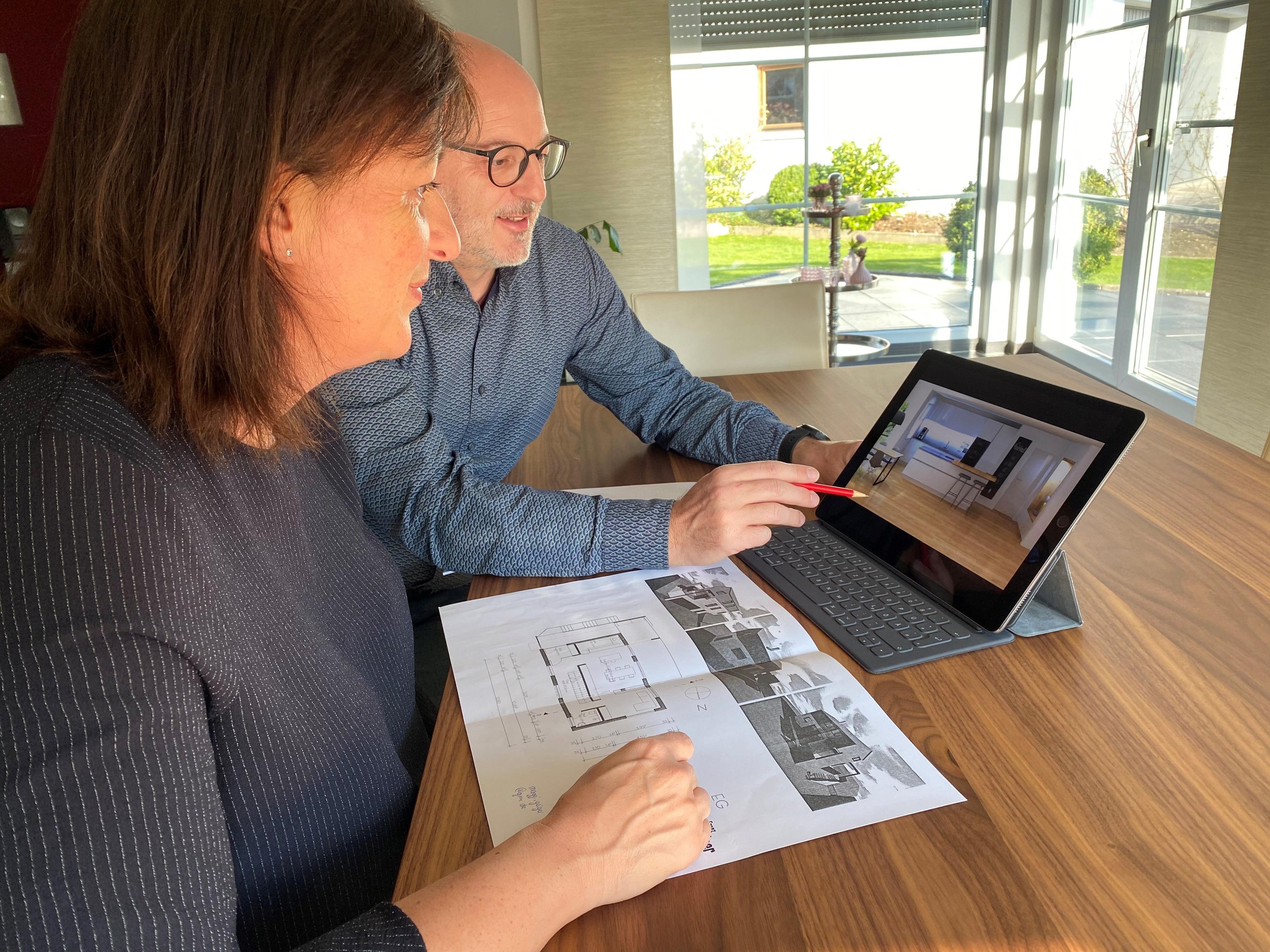 Mann und Frau betrachten gemeinsam eine 3D-Küchenvisualisierung auf einem Tablet, während Baupläne auf dem Holztisch ausgebreitet liegen.