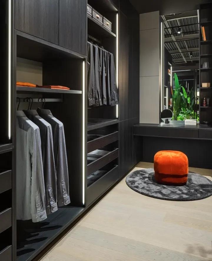 Stilvolles Ankleidezimmer in dunkler Holzoptik mit beleuchtetem Kleiderschrank, Spiegel und orangem Hocker auf rundem Teppich.