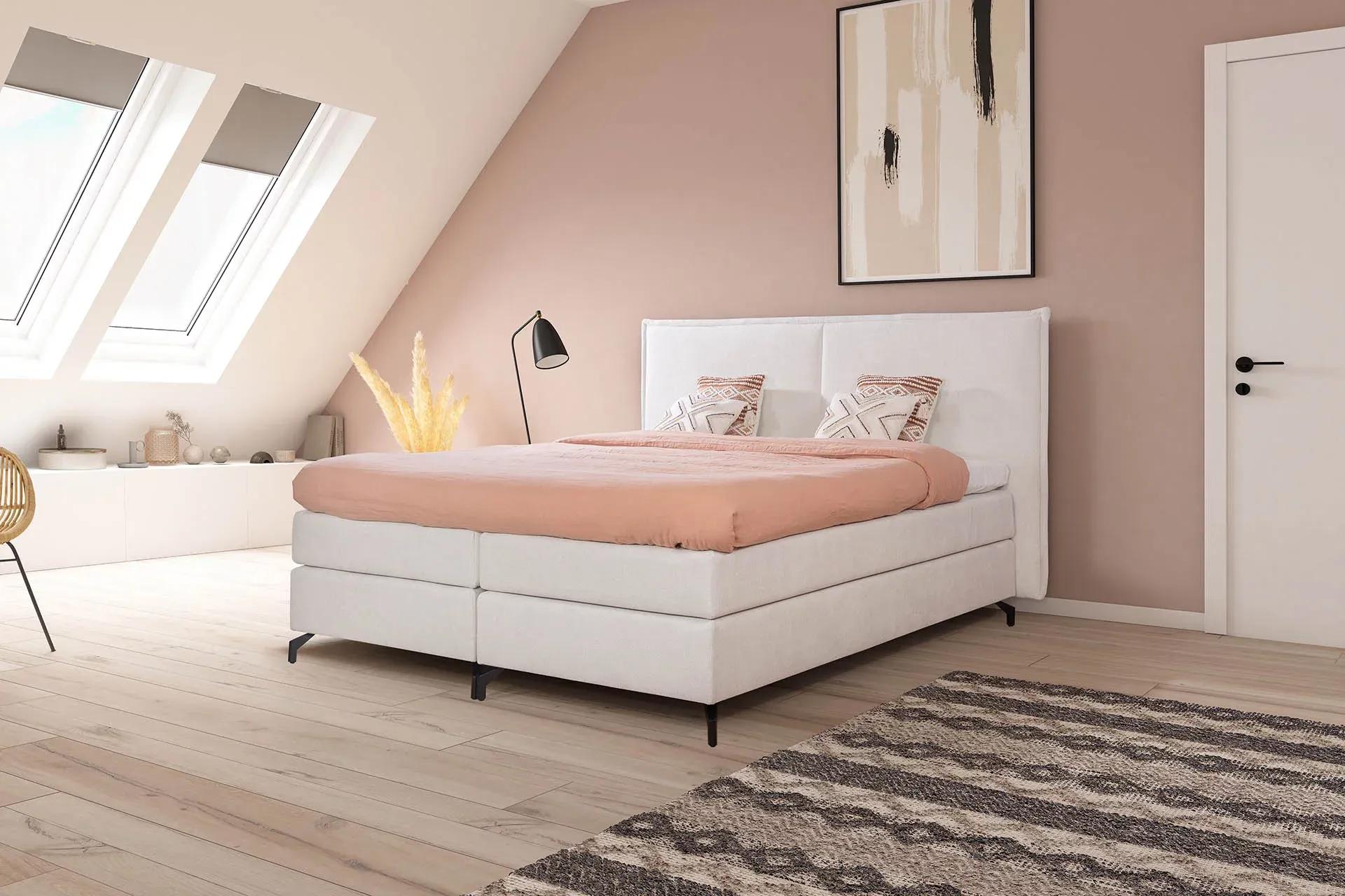 Modernes Boxspringbett in hellem Stoff mit hohem Kopfteil, roséfarbener Bettdecke und Dekokissen – gemütlich eingerichtet unter Dachschräge mit Oberlichtern.