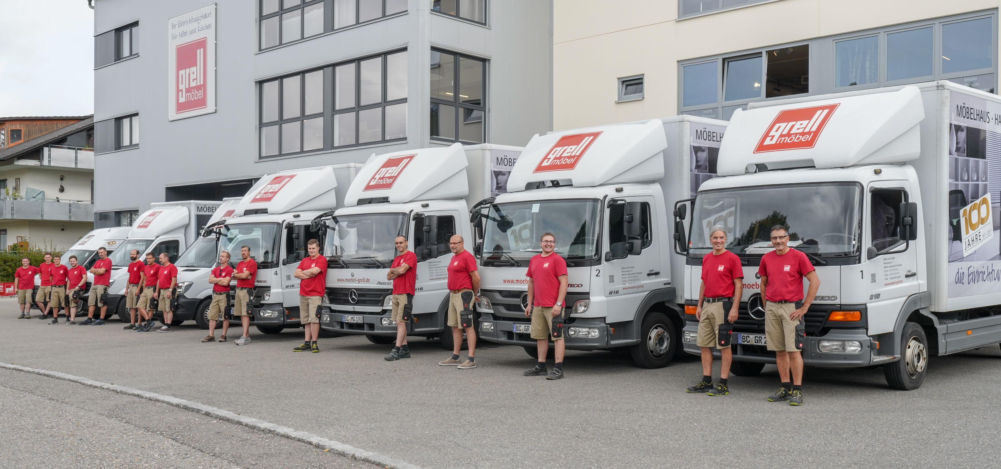 Firmenteam in roten T-Shirts steht vor einer Reihe weißer Lieferwagen mit "grell möbel" Logo vor dem Firmengebäude.