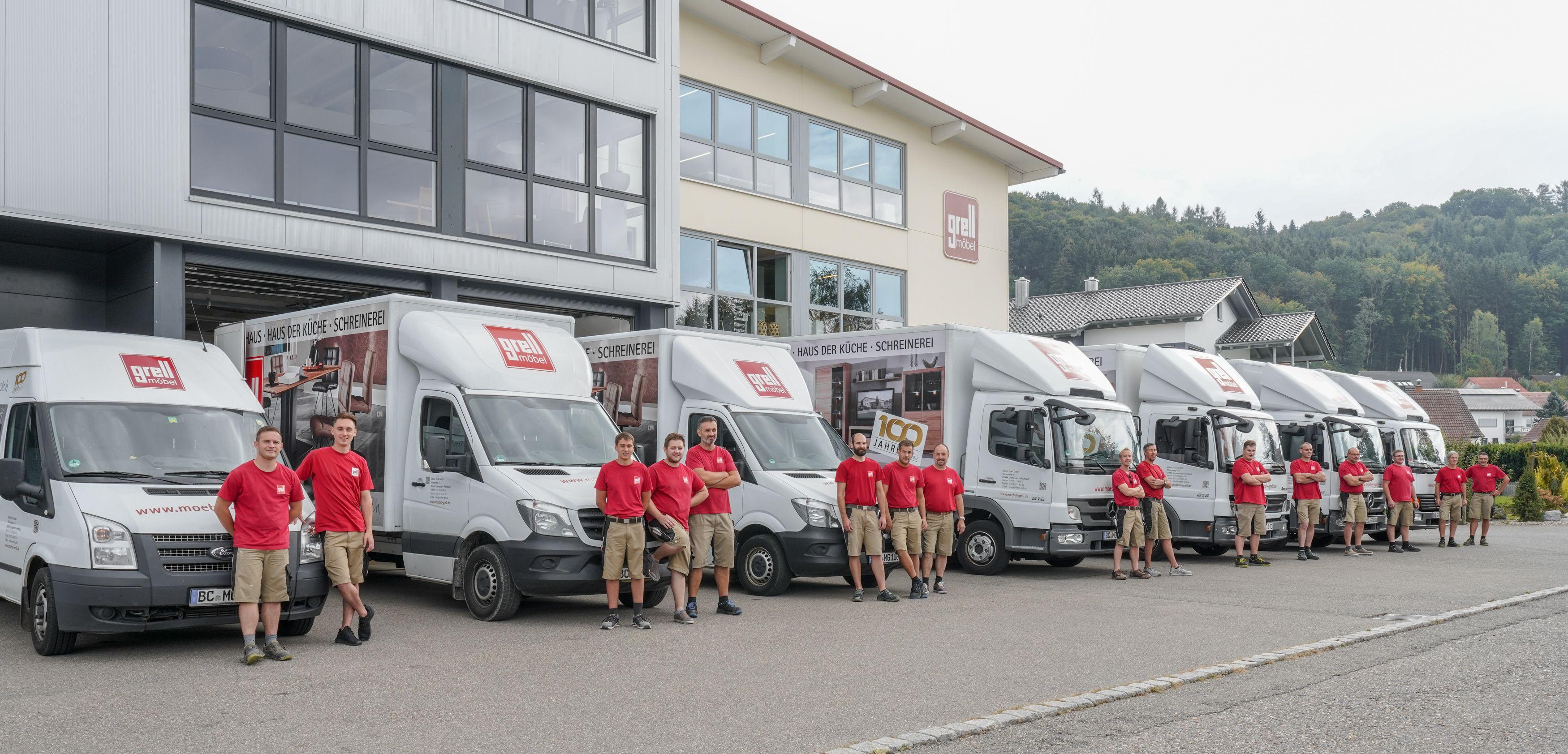 Team von Möbel Grell in roten T-Shirts steht vor einer Reihe weißer Lieferfahrzeuge vor dem Firmengebäude mit bewaldeter Landschaft im Hintergrund.