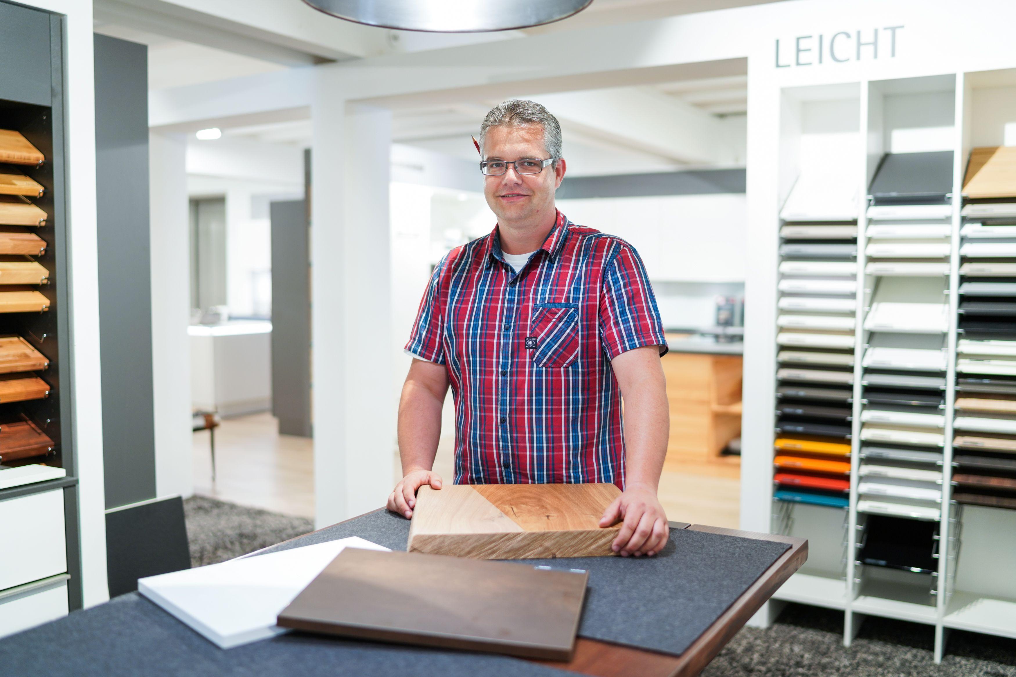 Mann mit Brille und kariertem Hemd präsentiert Materialproben an einem dunklen Tisch im Möbelstudio mit Ausstellungsregalen im Hintergrund.