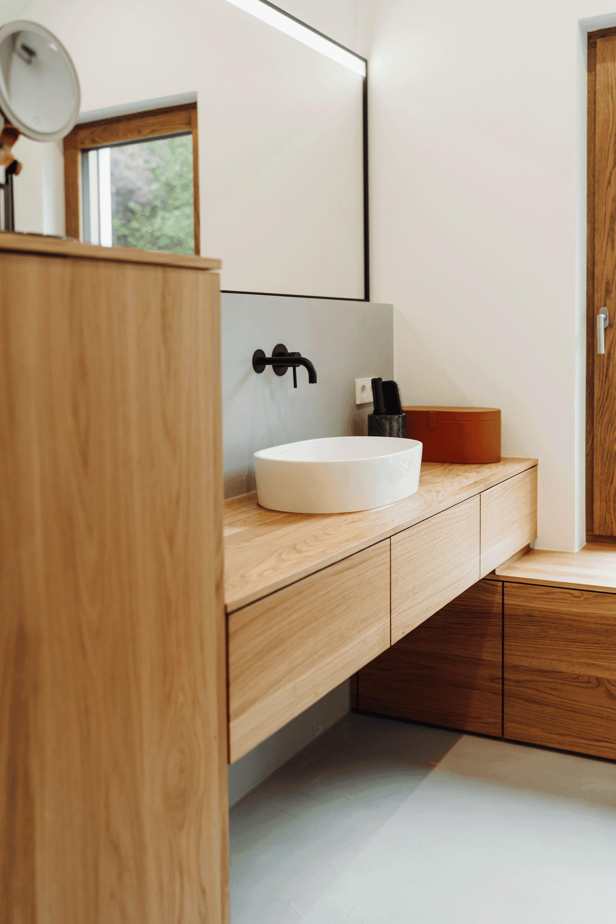 Badezimmer-Waschtisch aus Holz mit ovaler weißer Keramikschale, schwarzer Wandarmatur und Holzfenstern im Hintergrund.