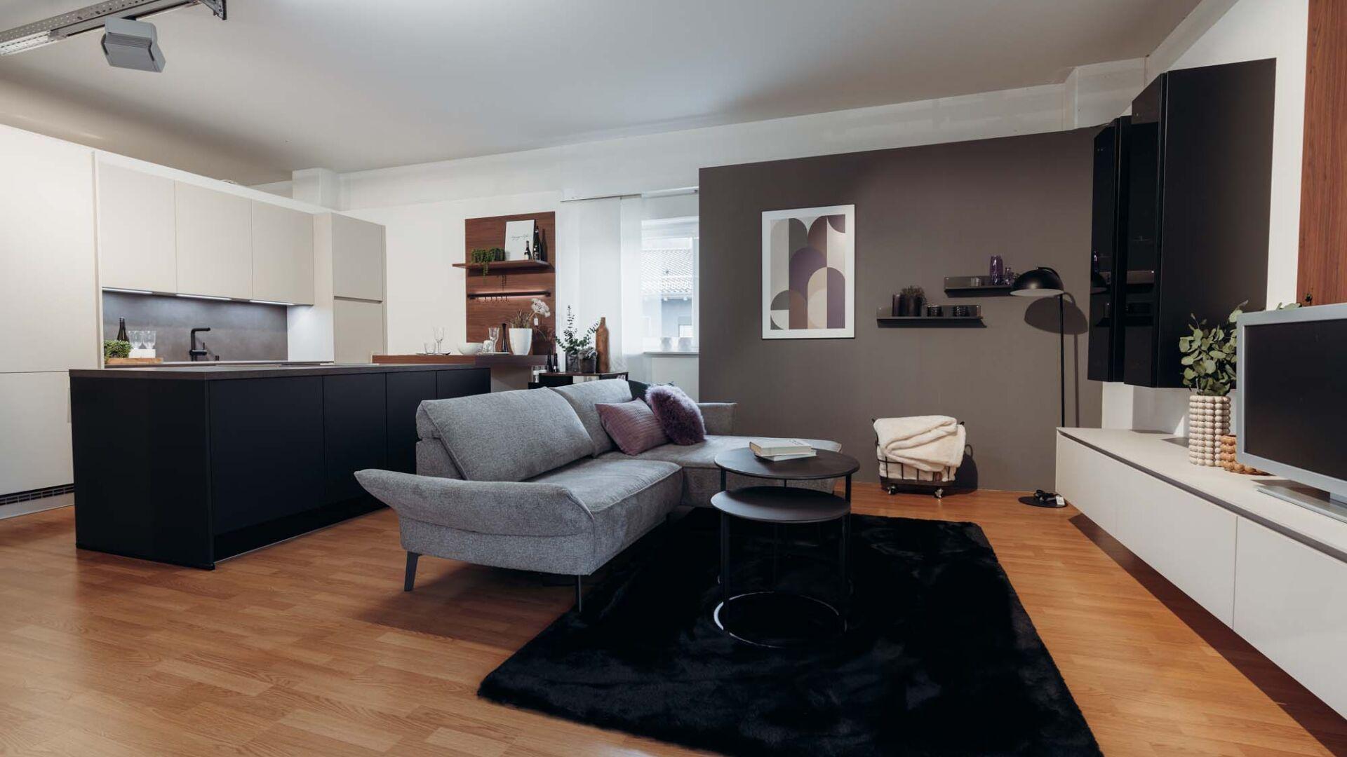 Offener Wohn-Küchenbereich mit grauem Sofa auf schwarzem Teppich, weißer Küche mit dunklen Unterschränken und modernem TV-Lowboard auf Parkettboden.