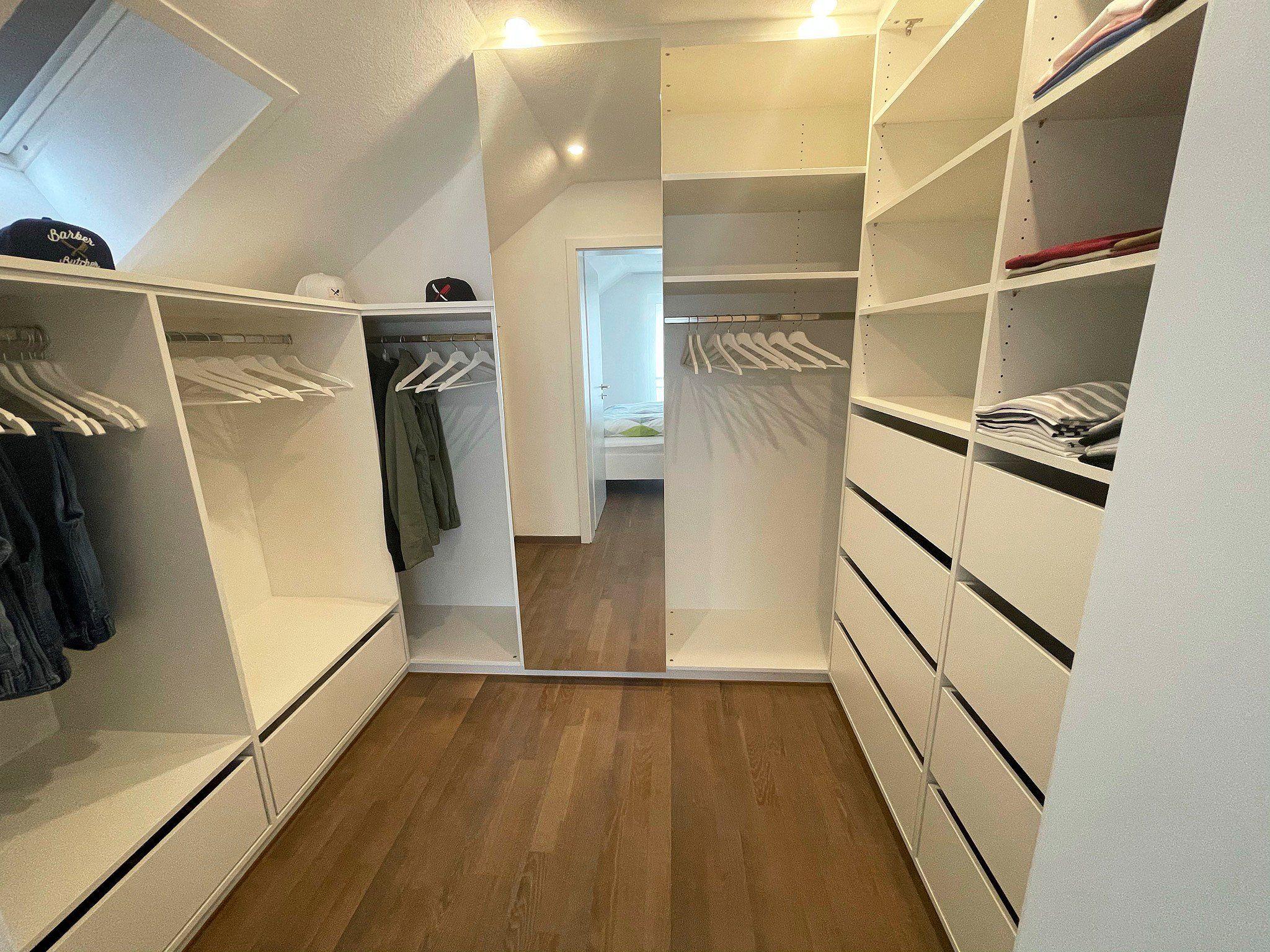 Maßgefertigtes Ankleidezimmer unter Dachschräge mit weißen Einbauten, offenen Kleiderfächern, Schubladen und integriertem Spiegel – gefertigt von Möbel Grell.