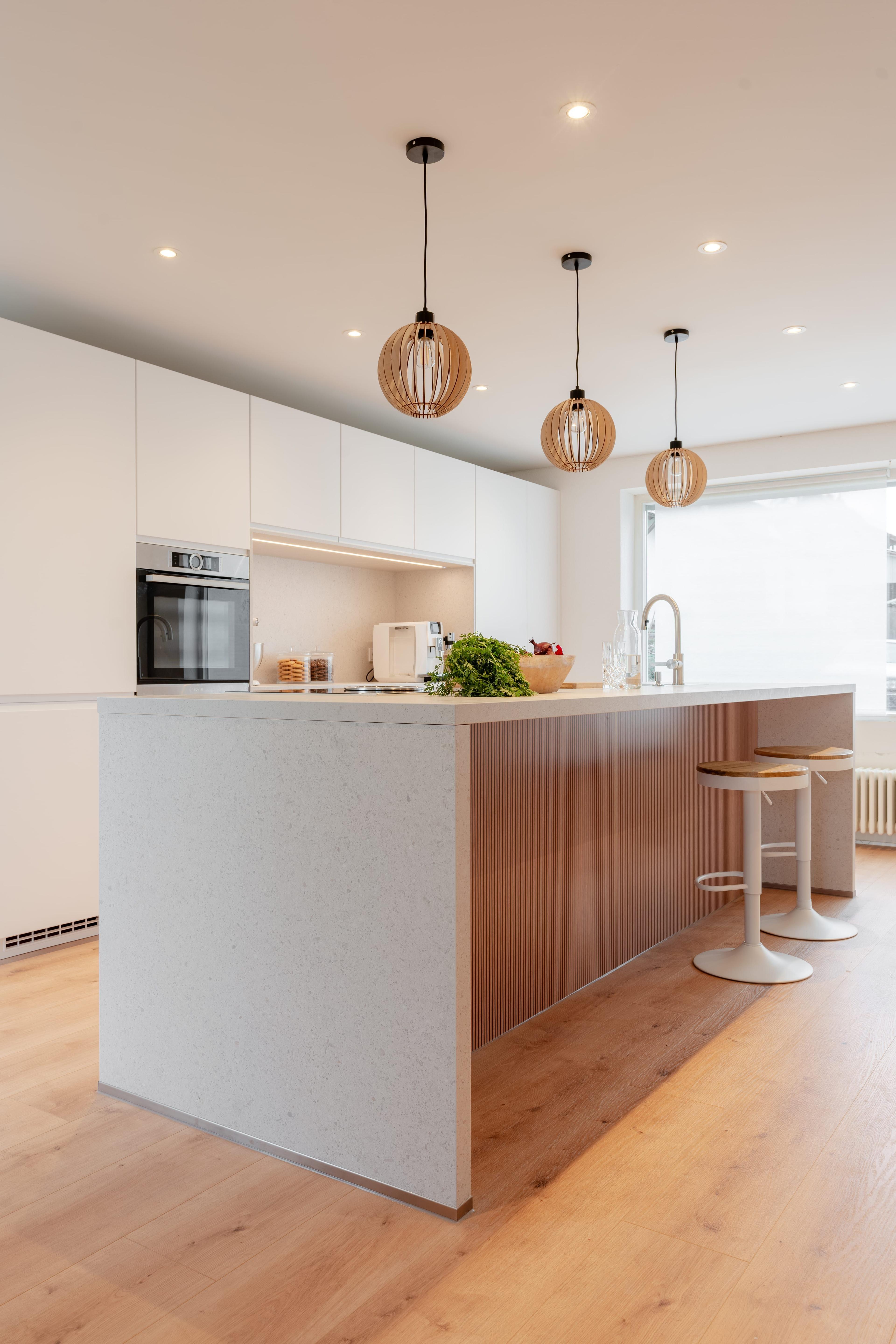 Moderne Küche mit weißen Oberschränken, Kücheninsel aus hellem Stein mit Holzverkleidung, drei Holz-Pendelleuchten und weißen Barhockern auf Parkettboden.