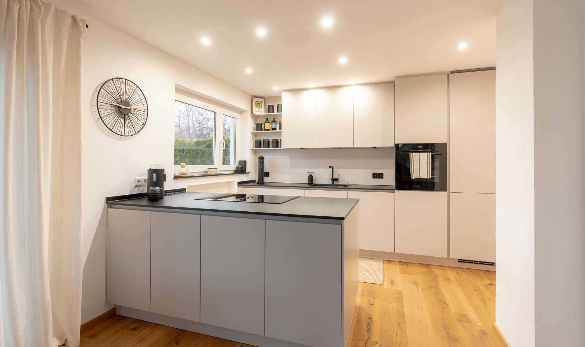 Offene Küche mit grauer Kochinsel, beigen Oberschränken, schwarzer Wanduhr und hellen Vorhängen am Fenster.