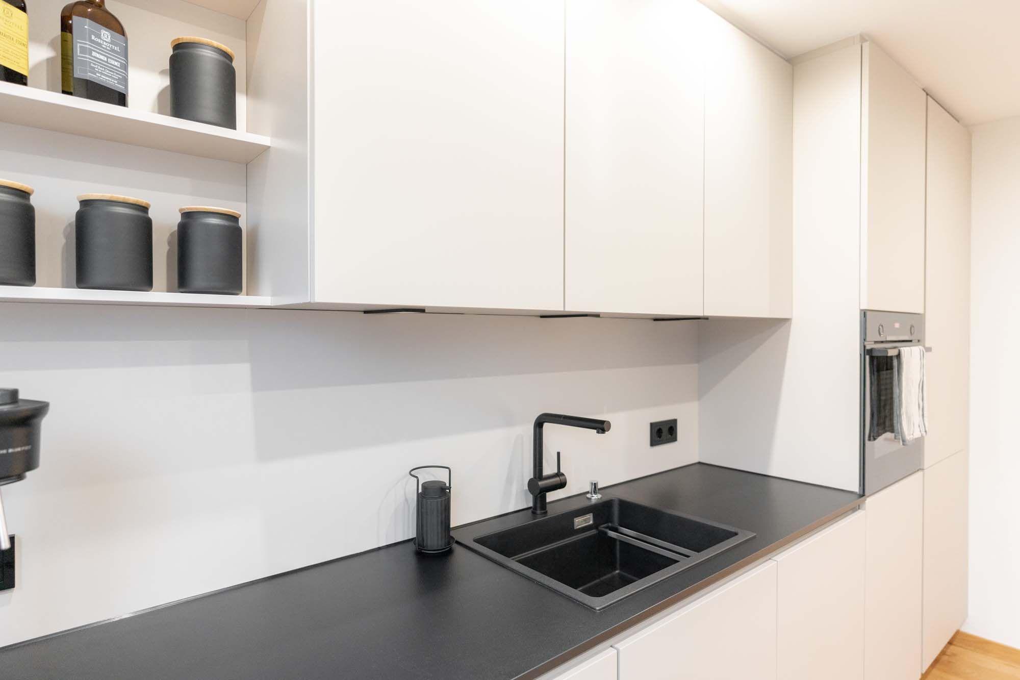 Moderne Küchenzeile mit grifflosen weißen Oberschränken, schwarzer Spüle und offenen Regalen mit Vorratsgläsern