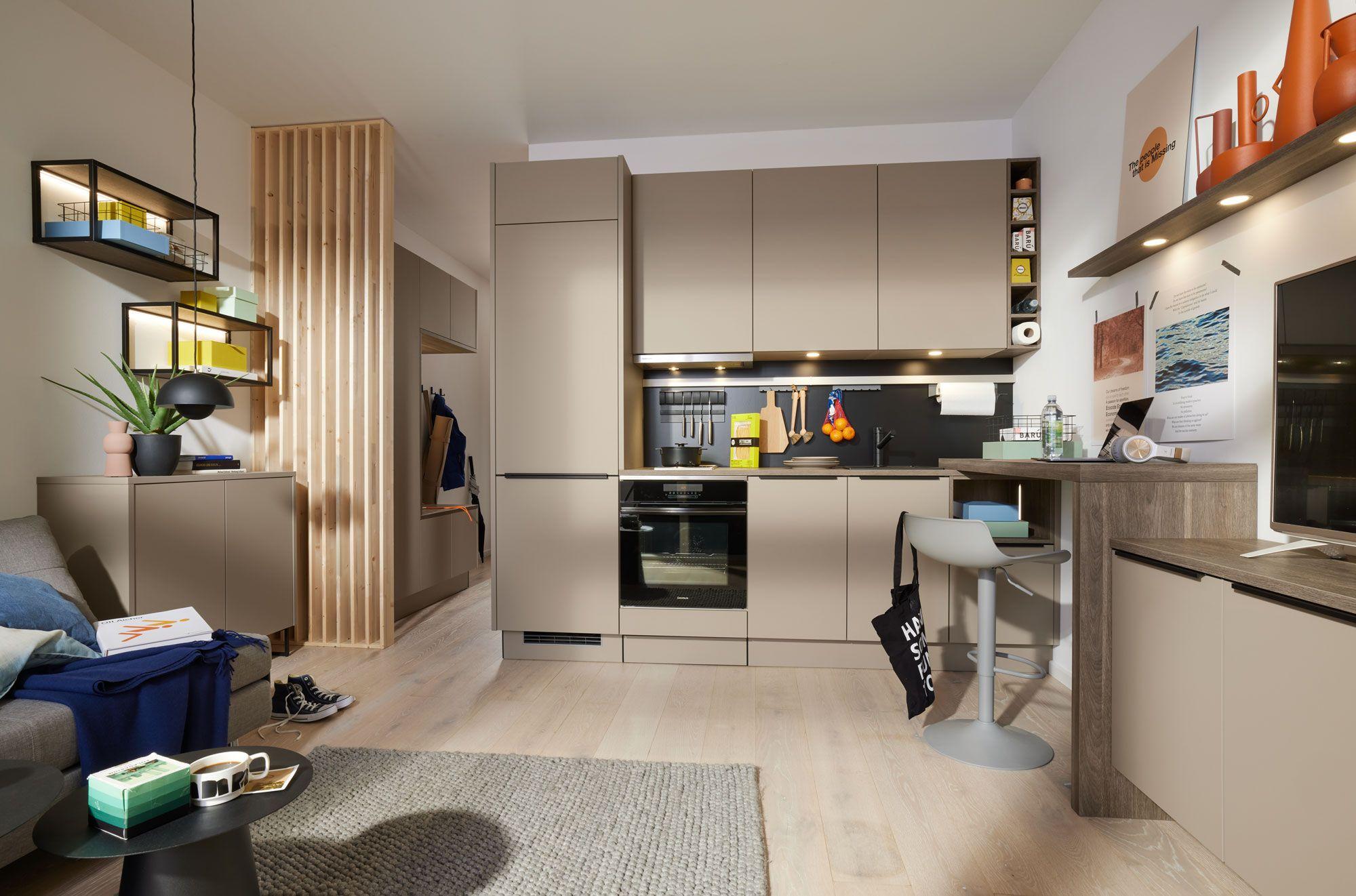 Moderne, minimalistische Küchenzeile in neutralem Beige mit eleganten Holzregalen und stilvollen Akzenten. Im Hintergrund sind ein Esstisch, ein Sofa und Dekor-Elemente sichtbar.