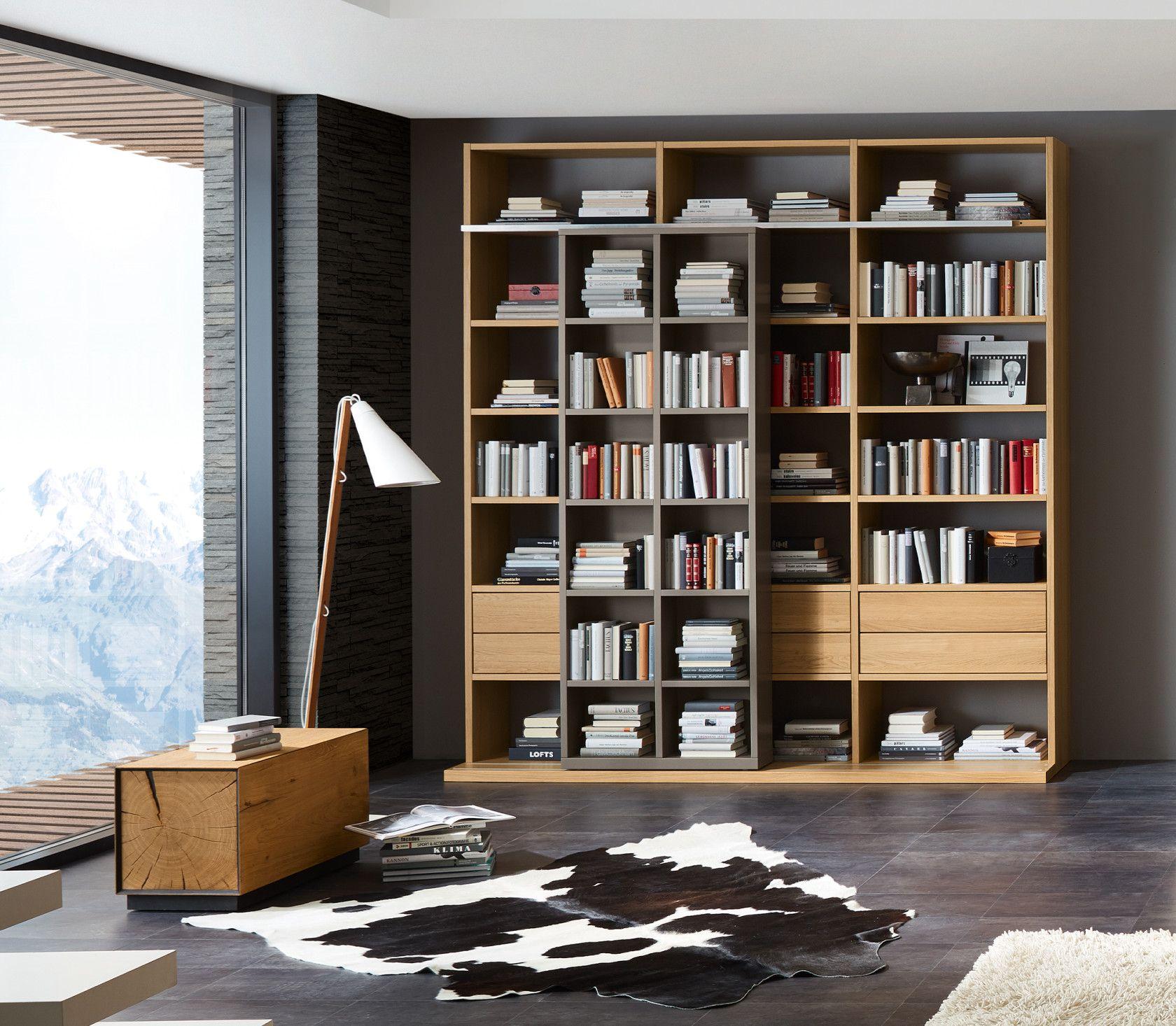 Großes Holzbücherregal mit integrierten Schubladen vor dunkler Wand, daneben eine Design-Stehlampe und Kuhfell-Teppich, Bergblick durch Panoramafenster.