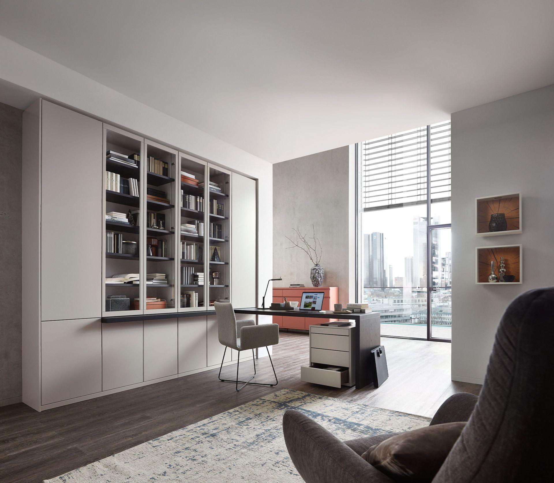 Elegantes Büro mit großer grauer Schrankwand, Glasvitrinen voller Bücher, modernem Schreibtisch und Stadtblick durch bodentiefe Fenster.