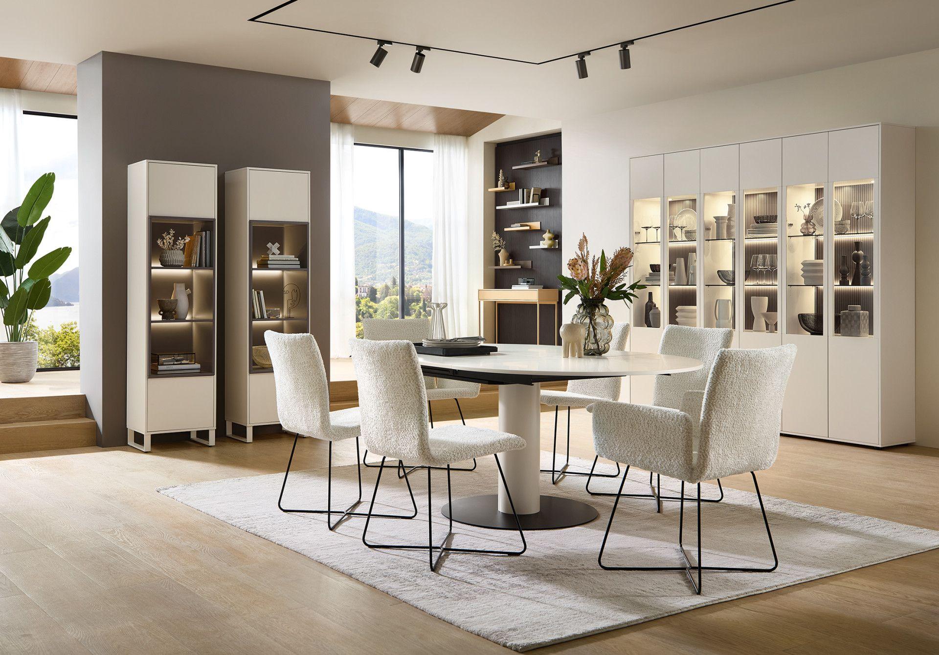 Modernes Esszimmer mit rundem Esstisch, weißen Stühlen mit Bouclé-Bezug, stilvollen Vitrinen und eleganter Dekoration in lichtdurchflutetem Raum.