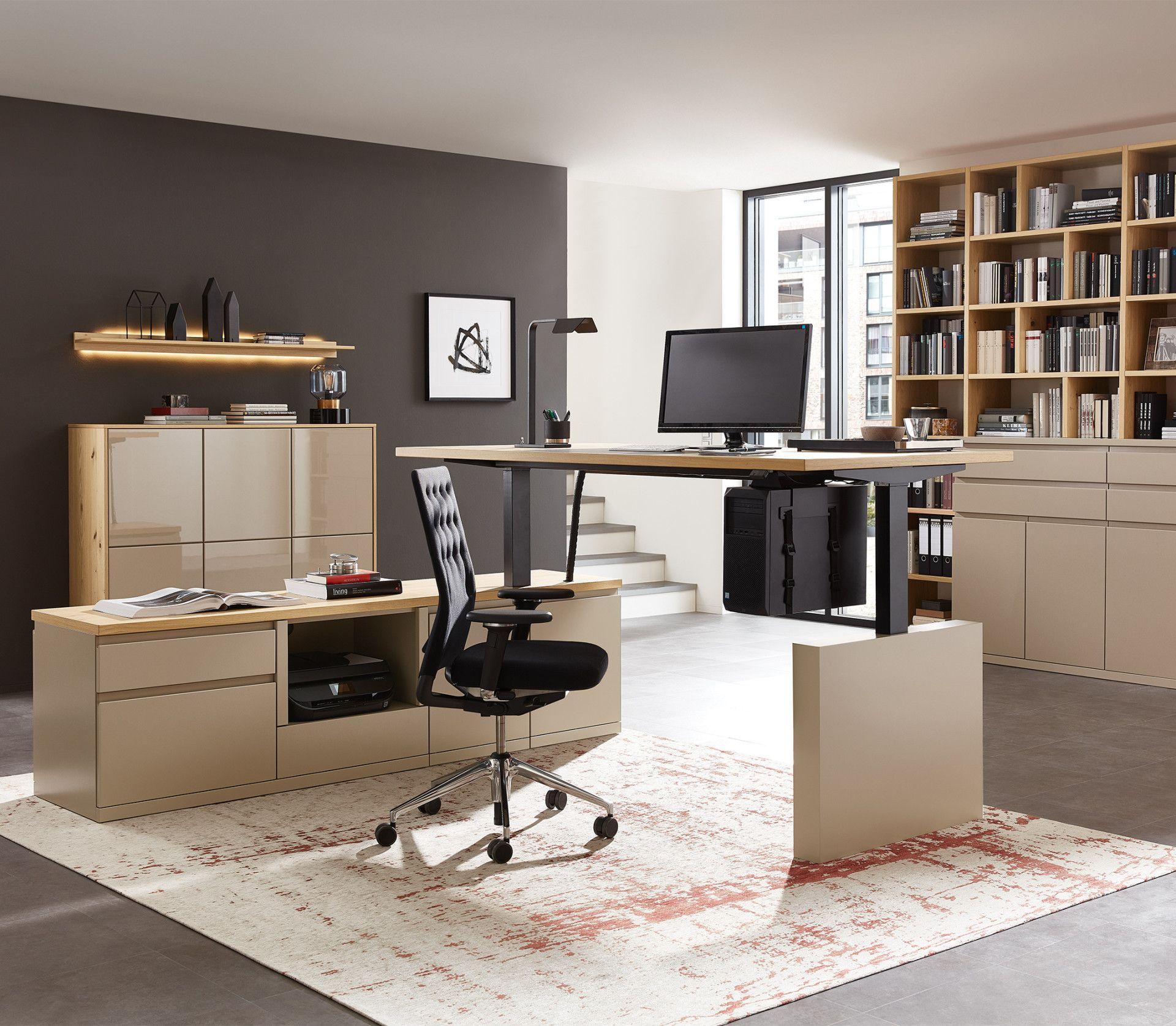 Elegantes Büro mit höhenverstellbarem Schreibtisch in Beige, schwarzem Bürostuhl und Holzbücherregal, graue Akzentwand und moderner Vintage-Teppich.