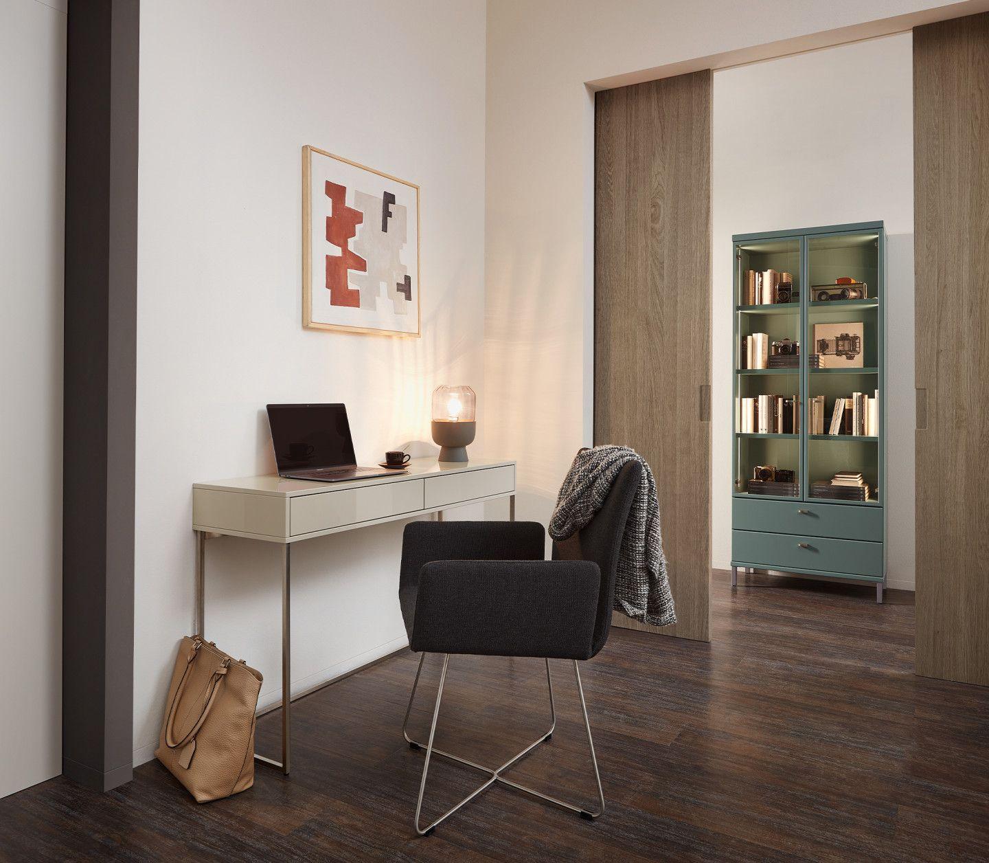 Minimalistischer Arbeitsplatz mit weißem Schreibtisch, dunklem Stuhl und Laptop, im Hintergrund grünes Bücherregal in Holznische und moderne Tischlampe.