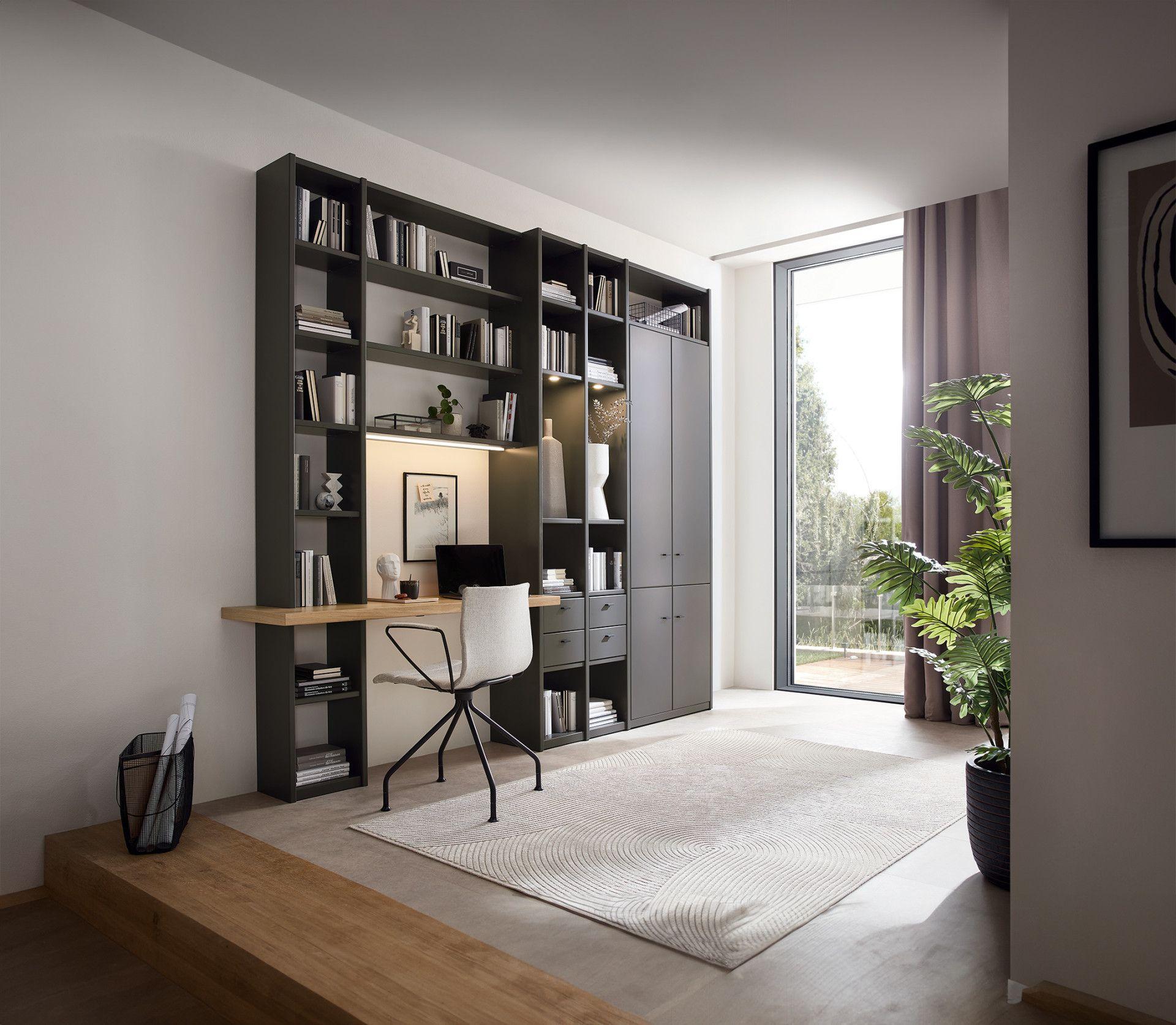 Elegante Bücherwand in Anthrazit mit integriertem Arbeitsplatz, weißem Designer-Stuhl und Schubladen, dazu ein strukturierter Teppich und große Zimmerpflanze.