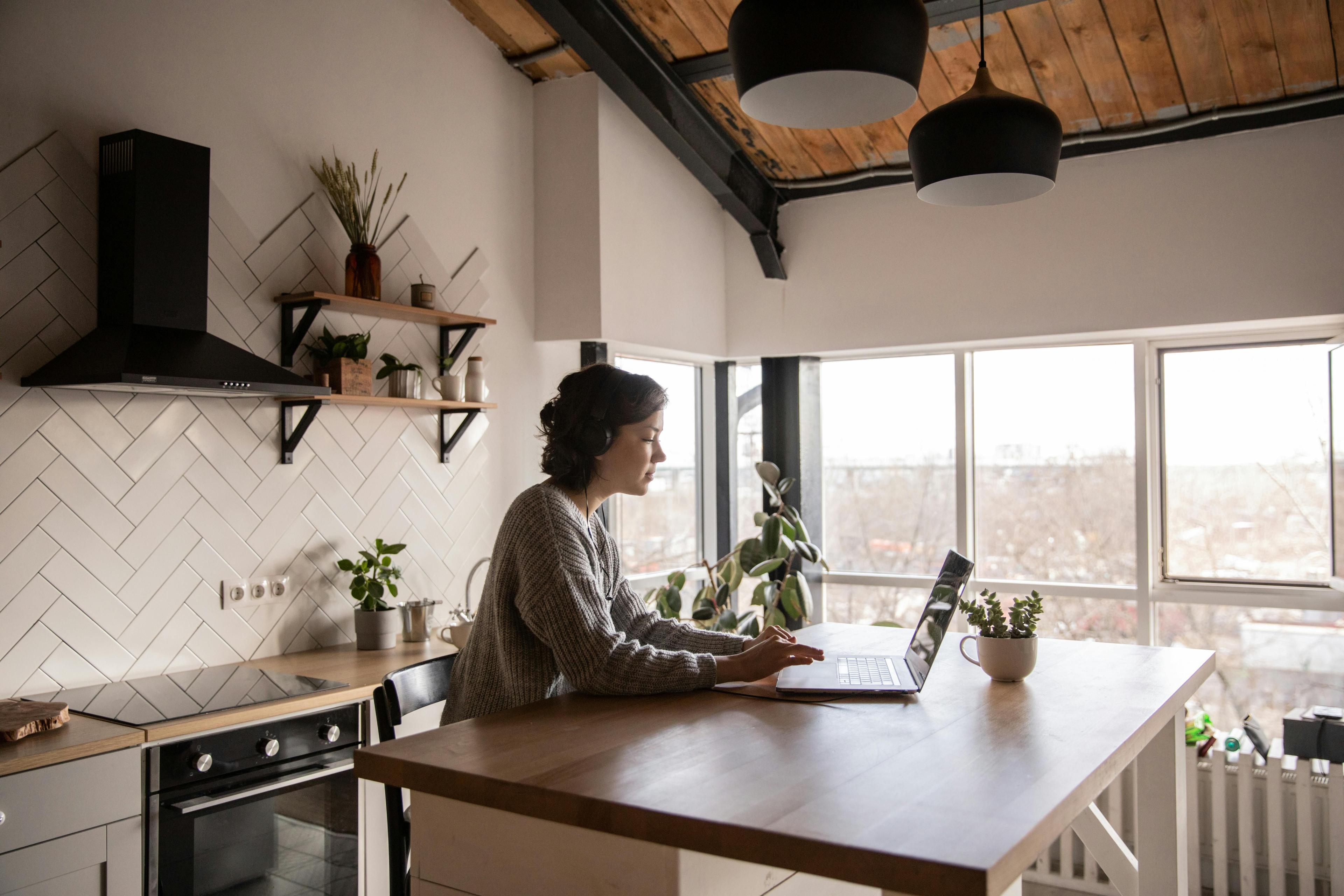 Frau arbeitet im Homeoffice an einem Laptop in einer modernen Küche mit Holzdecke, großen Fenstern und gemütlicher Einrichtung.