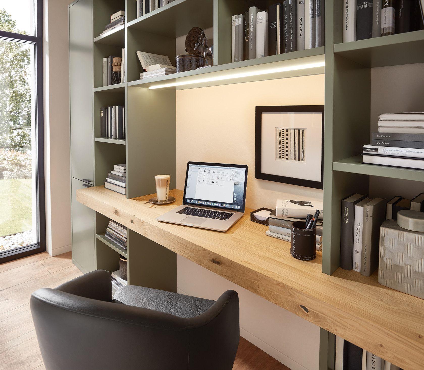 Nahaufnahme des Arbeitsplatzes mit Laptop auf Holzschreibtisch, umgeben von Bücherregalen in olivgrün, LED-Beleuchtung und Kaffeetasse.