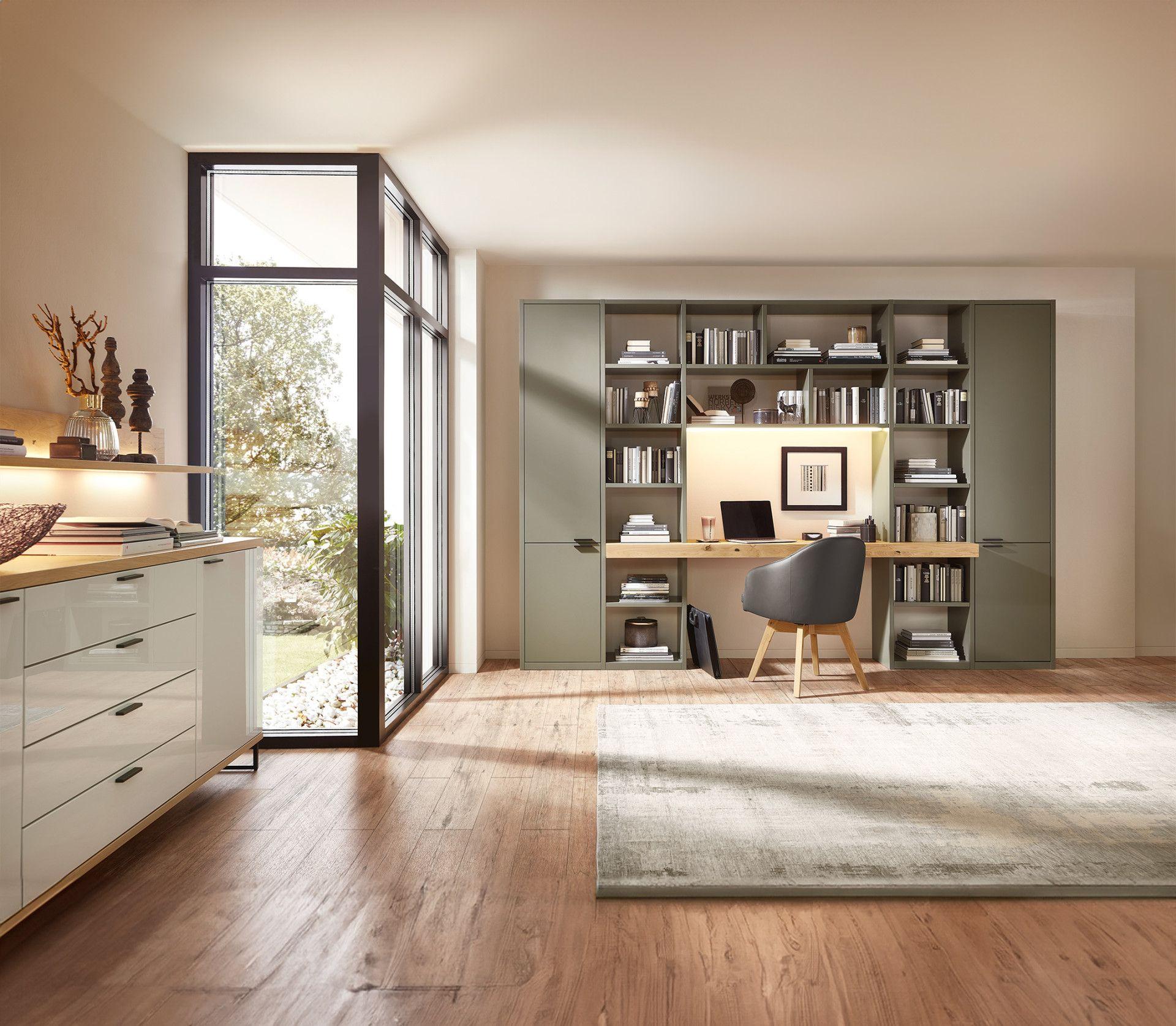 Modernes Home-Office mit grüner Bücherwand, integriertem Holzschreibtisch und schwarzem Drehstuhl, große Fensterfront mit Blick ins Grüne.