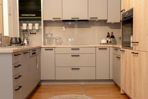 U-förmige Küche mit beigen Schränken und Holzoptik-Hochschrank rechts, Kücheninsel links mit Spüle. Runder geflochtener Teppich auf Parkett, industrielle Decke mit sichtbaren Leitungen.