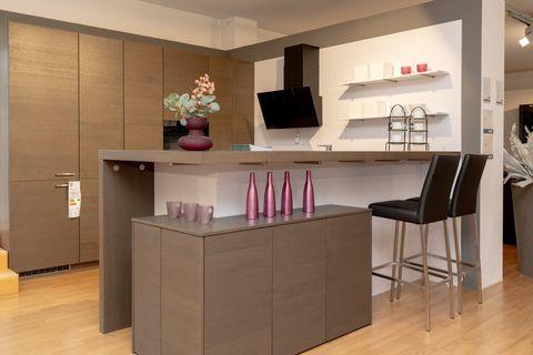 Moderne Küche mit holzfarbenen Oberschränken, grauer Kücheninsel mit rosa Dekorationsflaschen und schwarzen Barhockern. Weiße Wandregale und schwarze Dunstabzugshaube im Hintergrund.