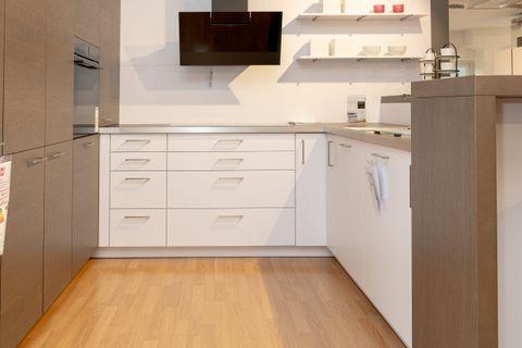 Moderne Küche mit holzfarbenen Oberschränken, grauer Kücheninsel mit rosa Dekorationsflaschen und schwarzen Barhockern. Weiße Wandregale und schwarze Dunstabzugshaube im Hintergrund.