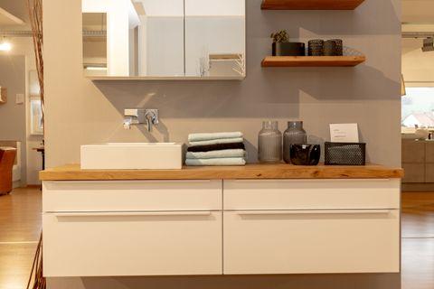 Modernes weißes Badmöbel-Set mit Eichenholz-Waschtischplatte, beleuchtetem Spiegelschrank und schwebenden Holzregalen in Badausstellung.