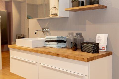 Modernes weißes Badmöbel-Set mit Eichenholz-Waschtischplatte, beleuchtetem Spiegelschrank und schwebenden Holzregalen in Badausstellung.