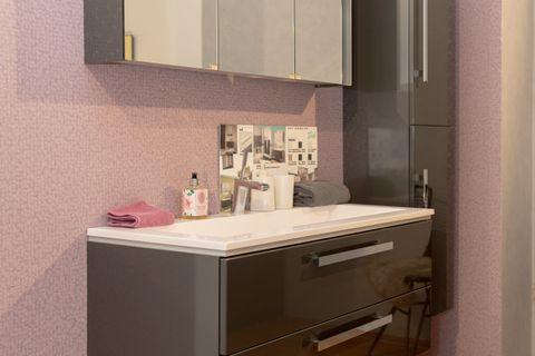 Modernes graues Badmöbel-Set mit beleuchtetem Spiegelschrank, schwebendem Waschtisch mit Schubladen und Hochschrank vor rosa Wand.