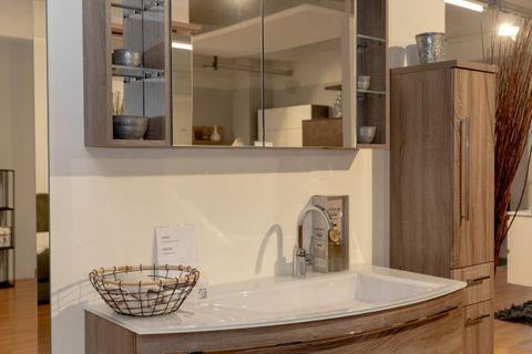 Moderner Waschtischunterschrank in Holzoptik mit weißem Waschbecken, Chromarmatur und dekorativem Drahtkorb in Badausstellung.