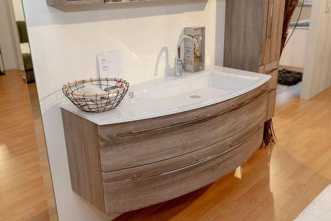 Moderner Waschtischunterschrank in Holzoptik mit weißem Waschbecken, Chromarmatur und dekorativem Drahtkorb in Badausstellung.