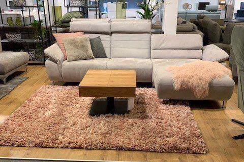 Hellgraue Eckcouch mit Chaiselongue und Holz-Couchtisch auf rosa Hochflor-Teppich in Möbelhaus-Showroom.