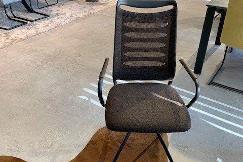 Moderner Esszimmerstuhl mit grauem Stoffbezug und perforierter Rückenlehne auf schwarzem Metallgestell in Möbelausstellung.