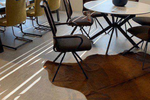 Moderner Esszimmerstuhl mit grauem Stoffbezug und perforierter Rückenlehne auf schwarzem Metallgestell in Möbelausstellung.