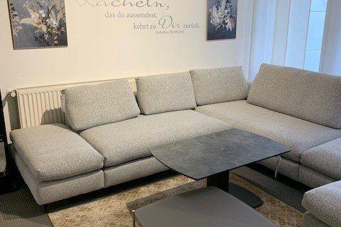 Große graue Stoff-Wohnlandschaft in U-Form mit schwarzem Couchtisch unter industrieller Holzbalkendecke.